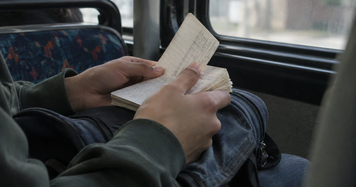 Close view of hands turning a handwritten flashcard above a backpack on a bus.