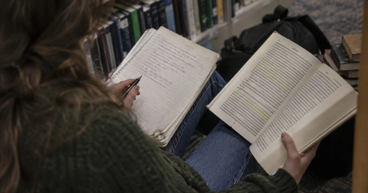 Hands hold an open handwritten notebook beside a highlighted book, with library shelves in the background.