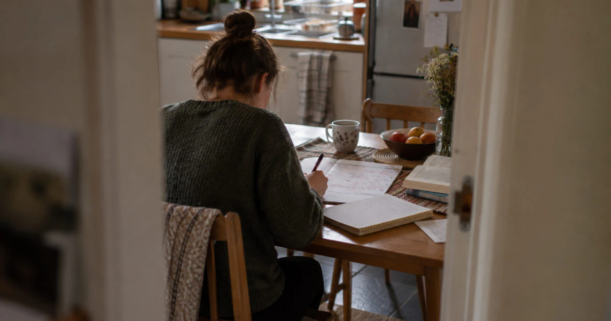 A student sits at a kitchen table with a handwritten calendar and an open notebook, viewed through a doorway.
