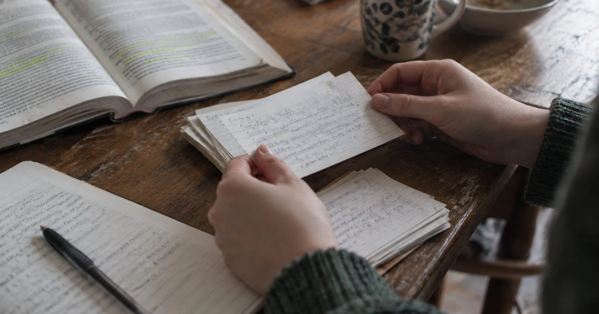 Hands holding a handwritten flashcard above a kitchen table beside a stack of cards and an open highlighted textbook.