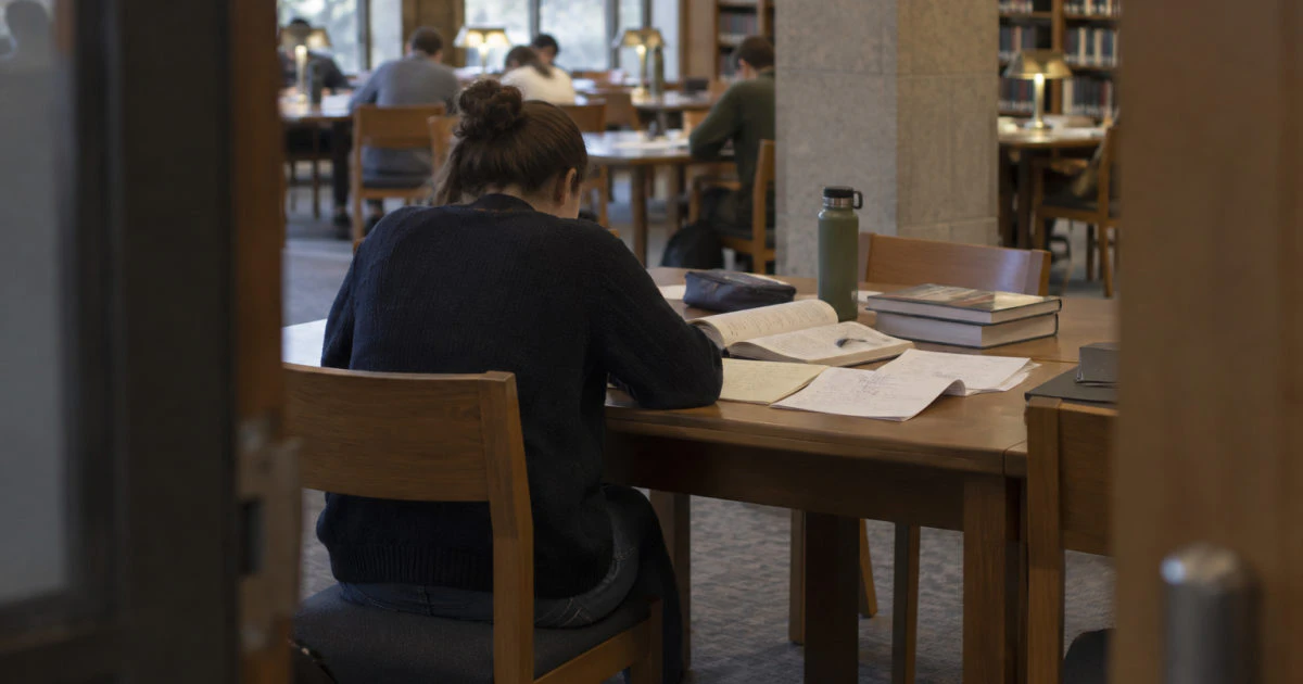A student seen from behind at a library table with annotated question sheets and an open handwritten notebook.