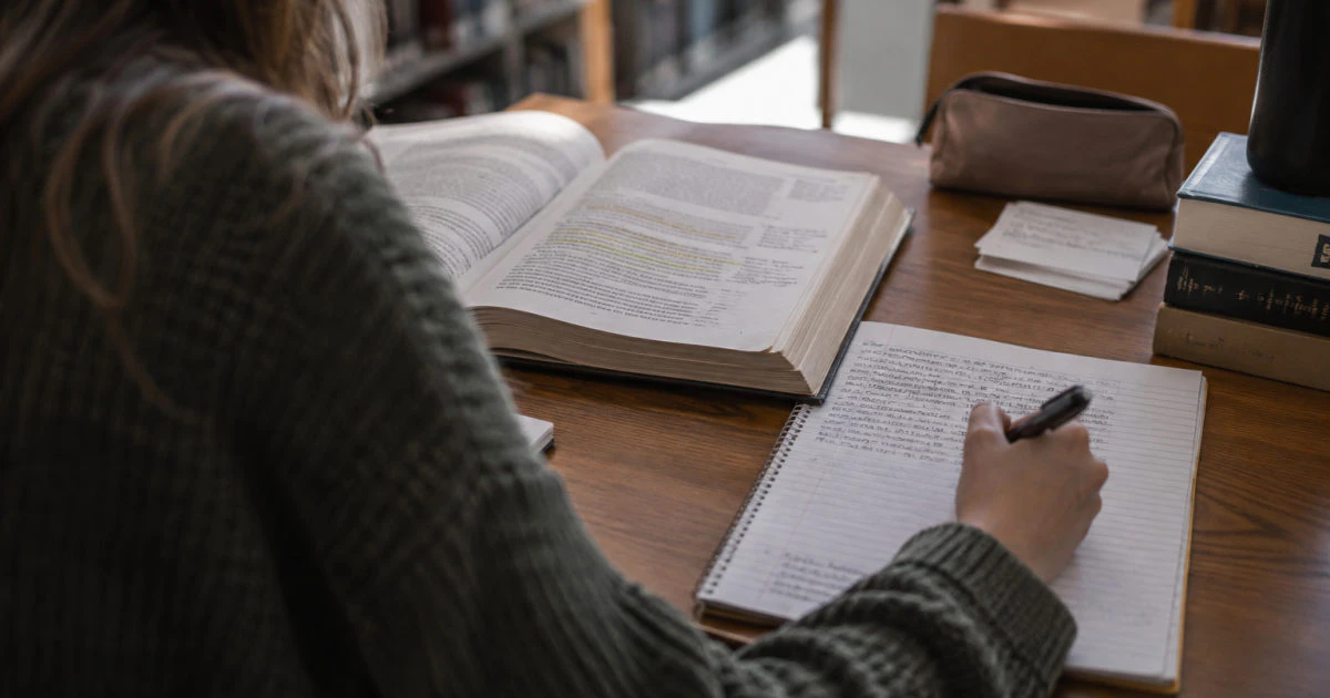 Over-the-shoulder view of a student writing in a part-filled notebook beside an open, highlighted textbook at a library desk.