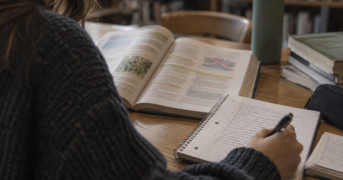 An open textbook with highlighted passages beside a notebook filled with handwritten notes on a library table.