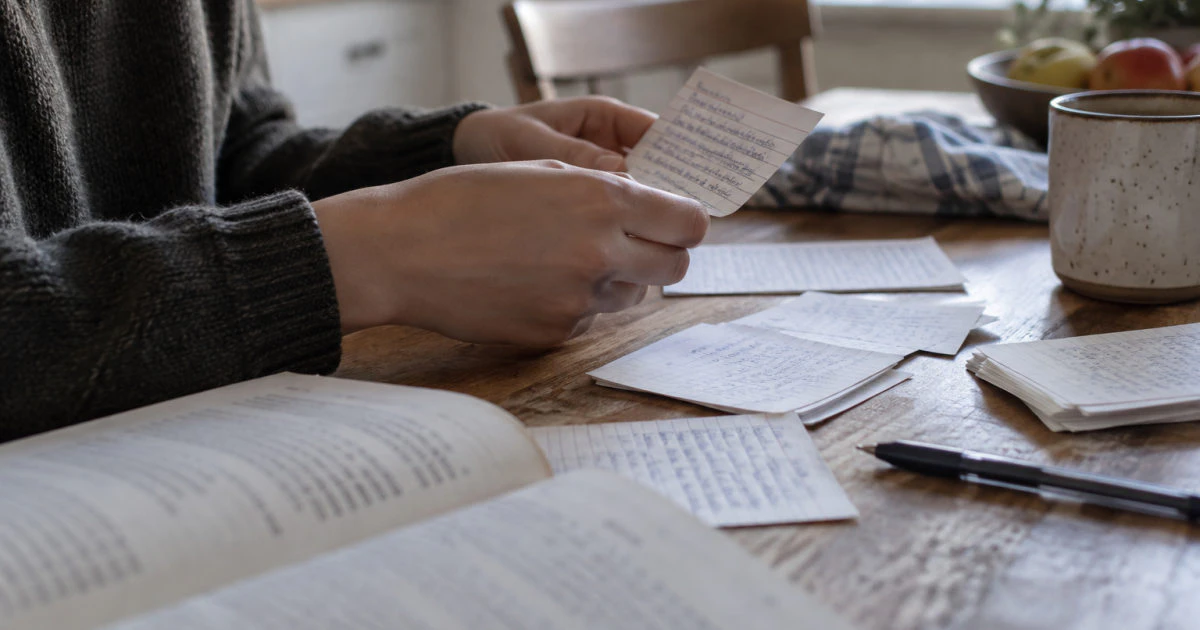 Hands holding a handwritten flashcard above several other written cards on a kitchen table.