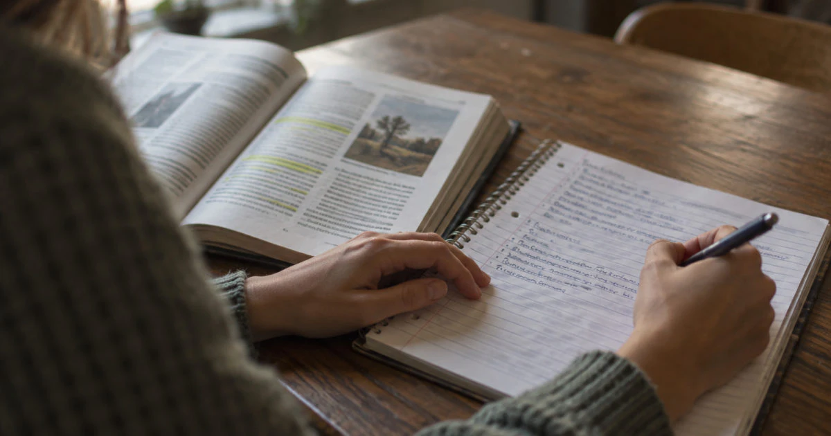 Hands writing on a partly filled notebook page beside an open, highlighted textbook on a kitchen table.