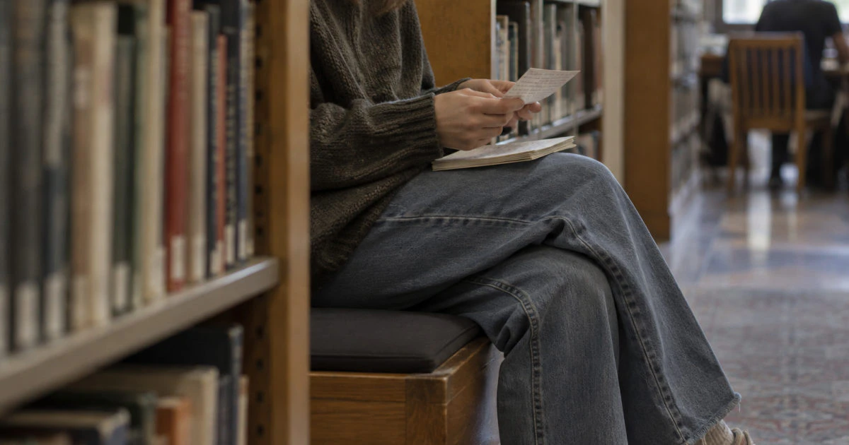 A student seated on a library bench holds a handwritten flashcard above a small stack of cards.